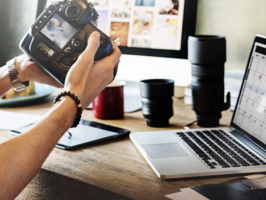 Hands holding a camera, with lenses, a tablet, and a laptop in background.